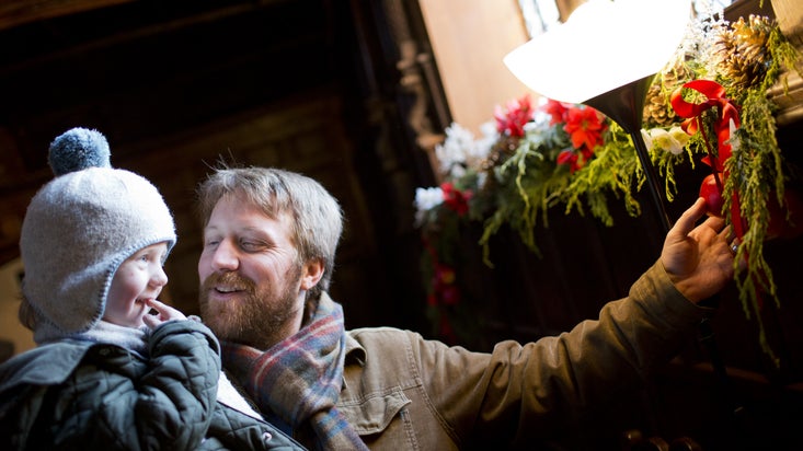A man holds a young child near a window decorated with Christmas ornamentation. The child wears a knit hat and quilted coat; the man wears a corduroy jacket and plaid scarf. Both smile.
